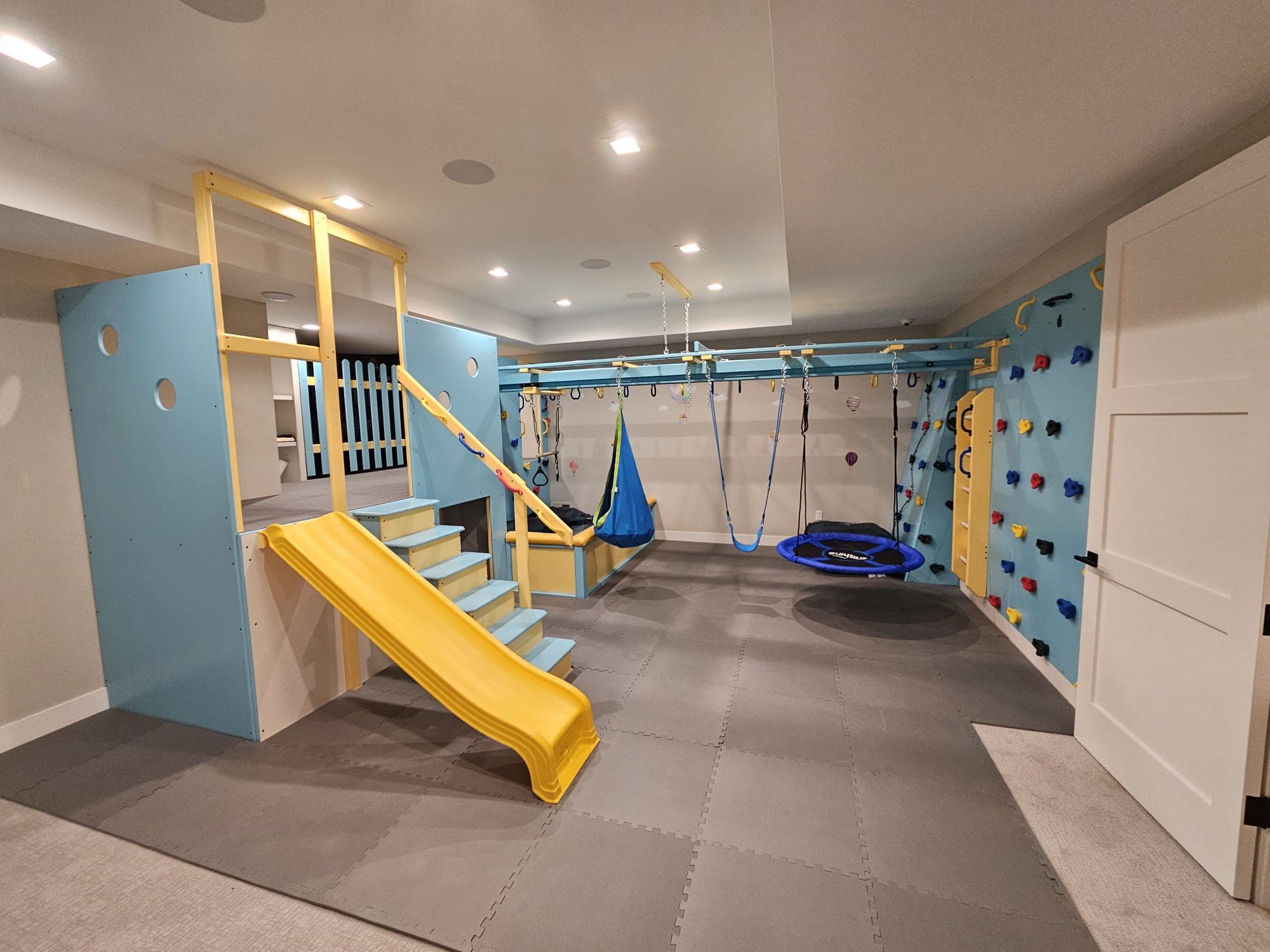 Custom home sensory gym with yellow slide, climbing wall, and swings in a finished basement
